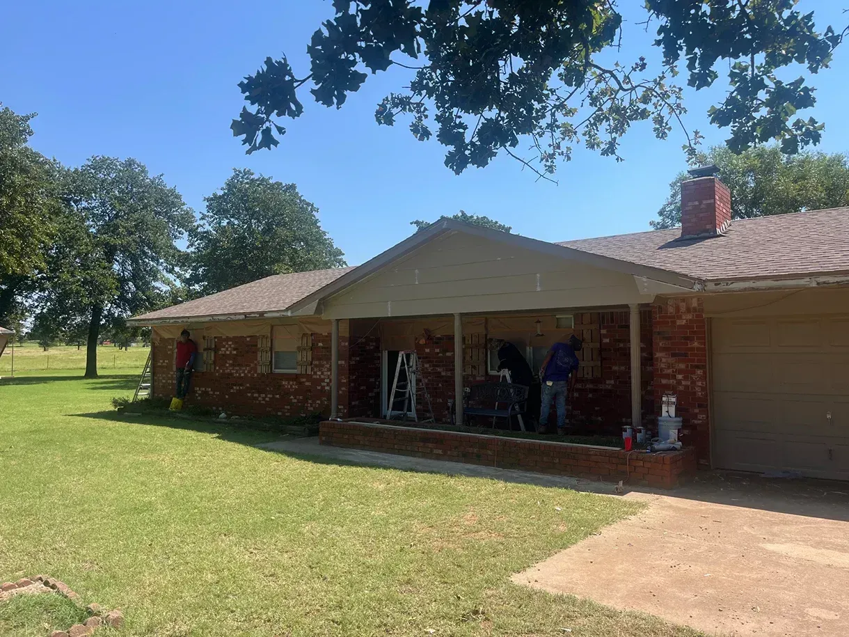 Brick house with porch being worked on, two people, ladder, grass lawn, clear sky.