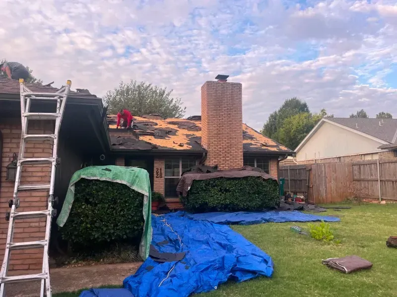 Roofing work in progress on a residential home with a damaged roof, ladder, and tarps.