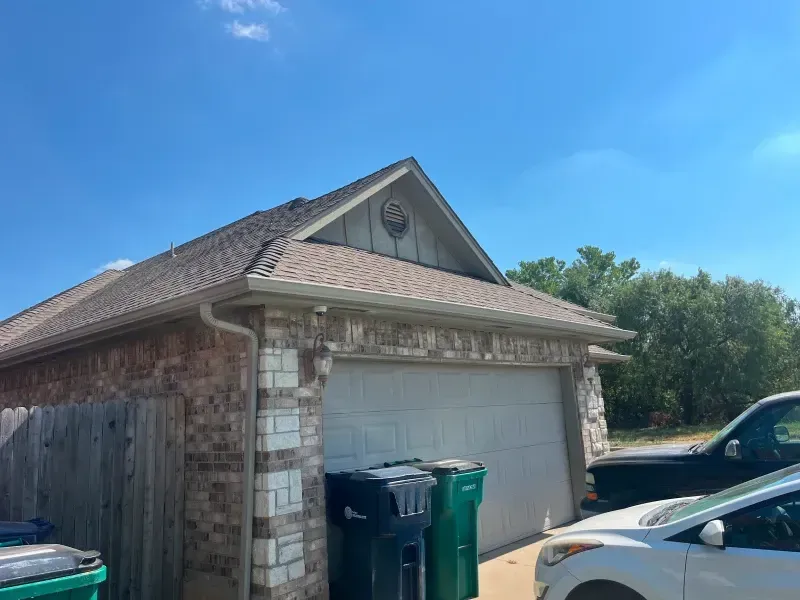 Garage with brown roof, tan gutters, brick and stone exterior, two trash cans in front, blue sky.