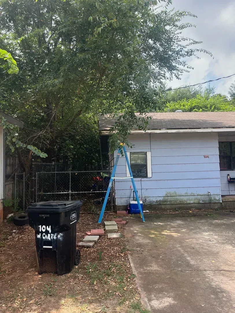 Blue house with ladder, overgrown tree, black trash bin, and cracked concrete driveway.