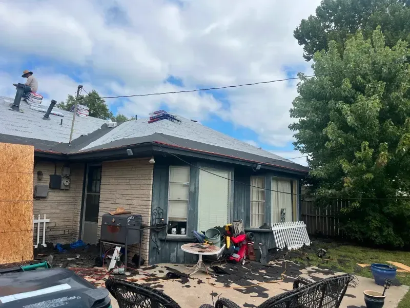 Roofer on a house roof replacing shingles. Yard debris and outdoor furniture are visible. Cloudy sky.