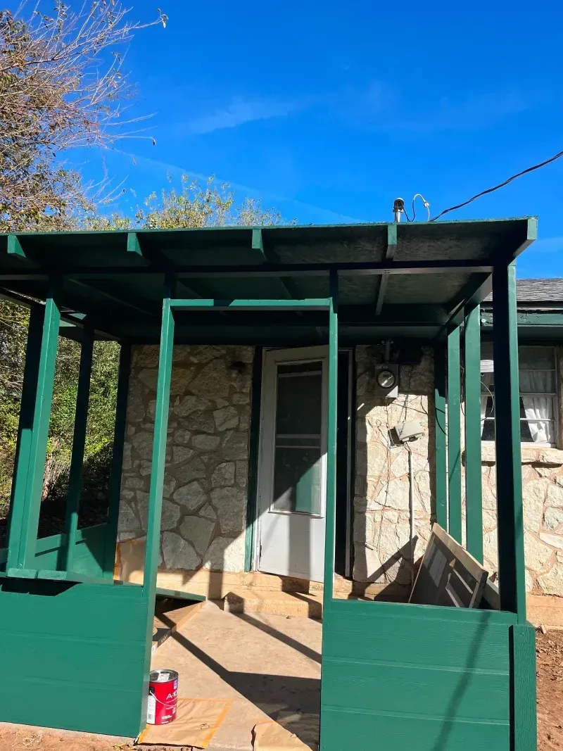 Green-painted porch of a stone building, white door, bright blue sky, paint can on ground.