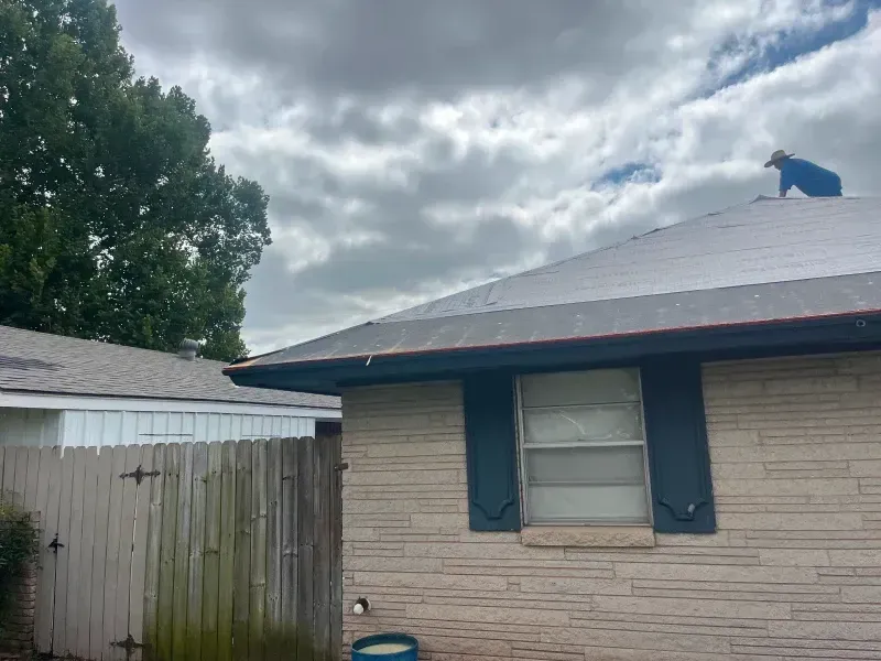A person on a roof working. Cloudy sky over a brick home and fence.