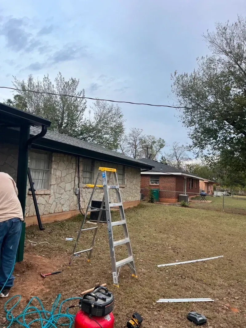 A person working on a house exterior. A-frame ladder, air compressor, tools, and house with a dark roof.