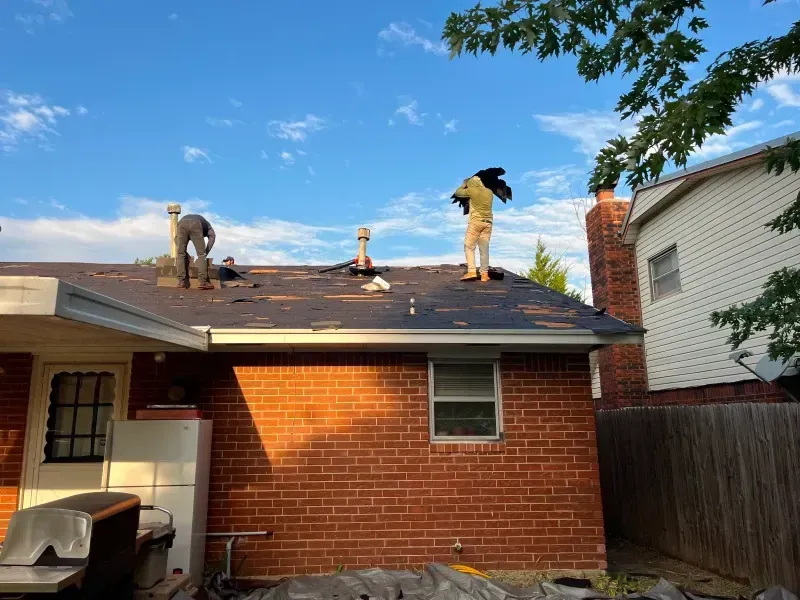 Two workers on a roof, removing shingles. Brick house with a blue sky.