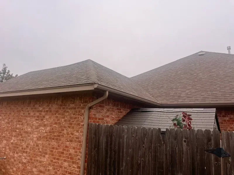 Brick house with brown shingle roof, brown gutters, and wooden fence on a cloudy day.