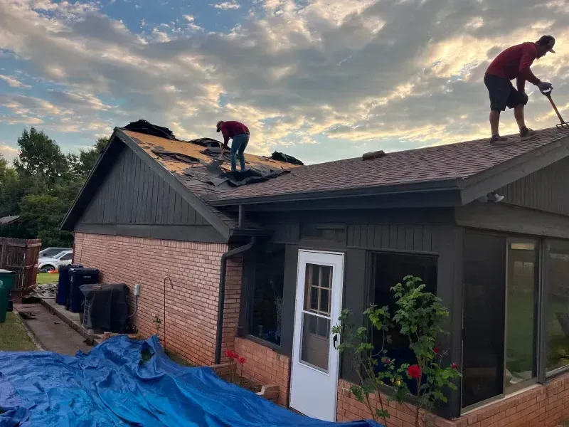 Two workers removing shingles from a house roof on a cloudy day.