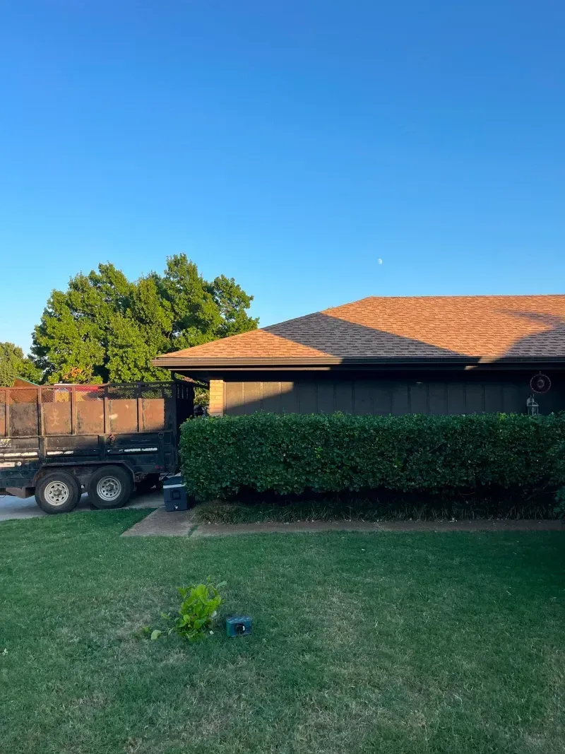 A house with brown roof, green hedge, and a trailer under a blue sky.
