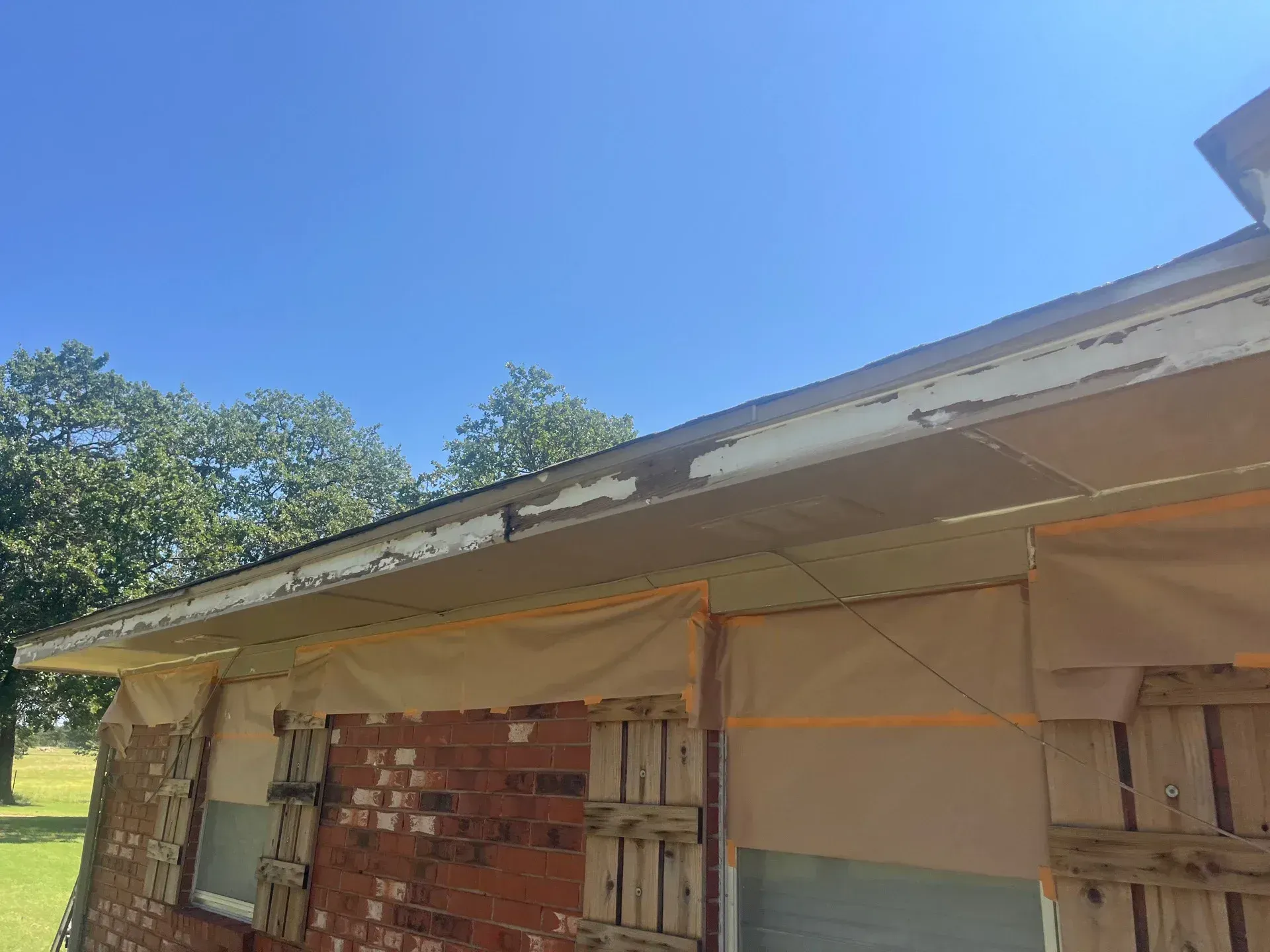 Exterior of building with peeling paint on eaves, covered brick, and protective sheeting; clear blue sky.