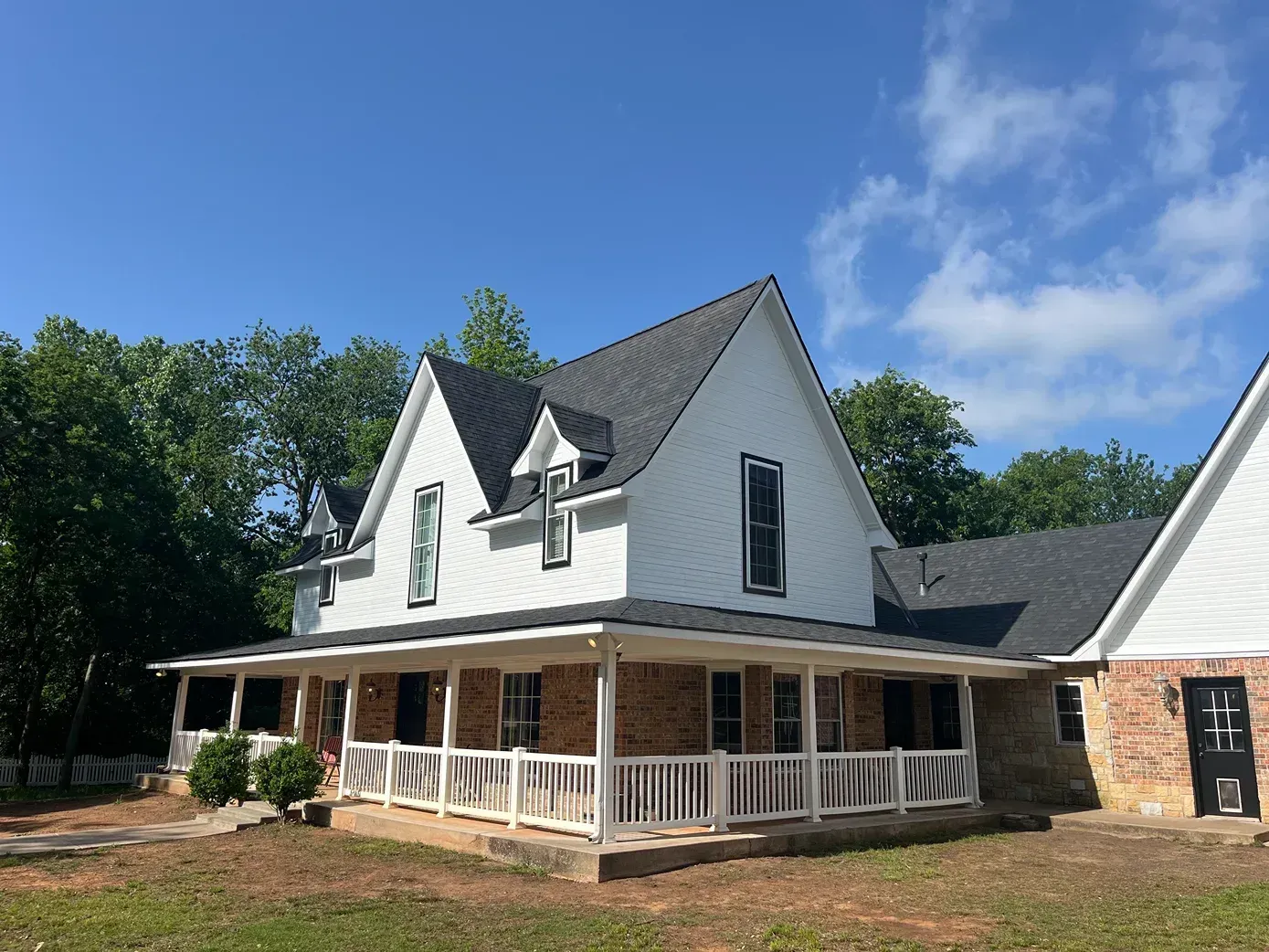 White farmhouse with black roof and a wrap-around porch, brick base, and a clear blue sky.