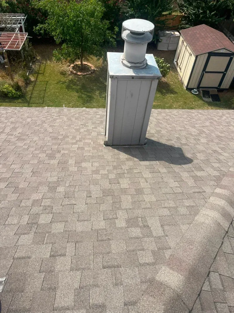 Chimney on a shingled roof casting a shadow in sunlight, with a shed and trees in the background.