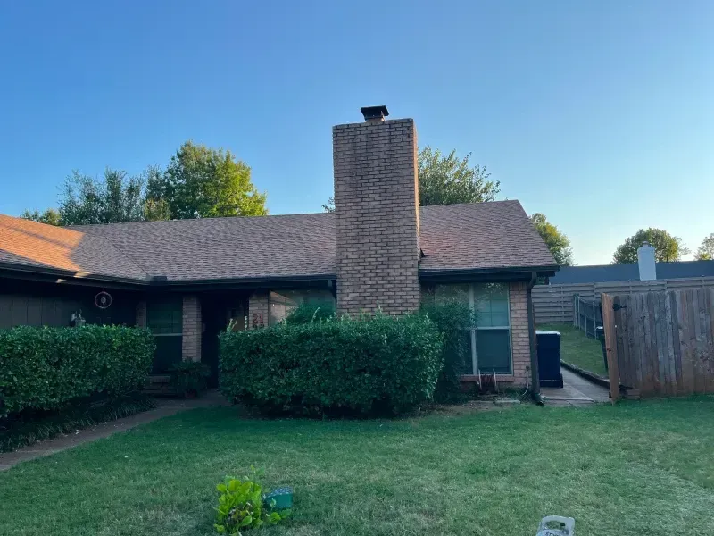 A brick chimney on a brown-roofed house with green bushes and a lawn under a blue sky.