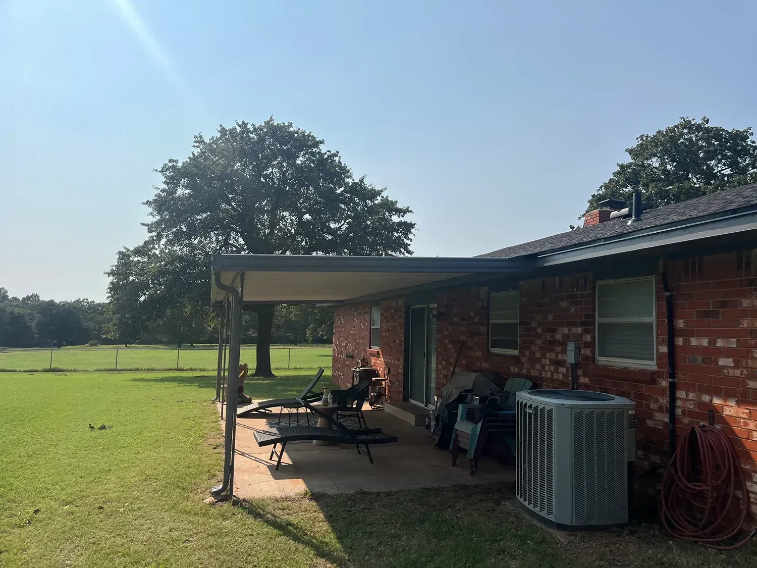 Backyard patio with a covered seating area attached to a brick house. Sunny day, green grass, and trees.