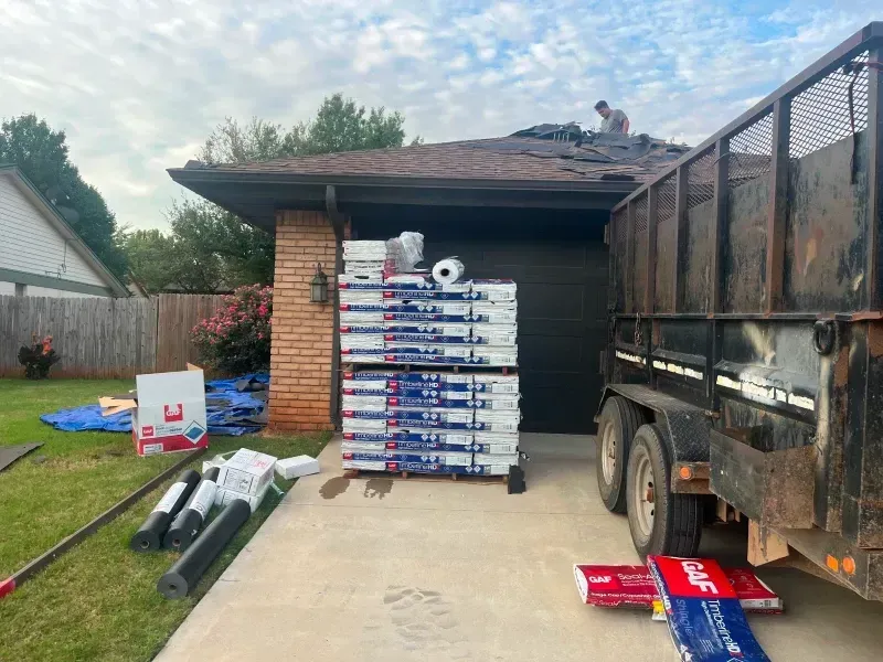 shingles stacked on driveway near a house. A worker is on the roof removing old shingles.