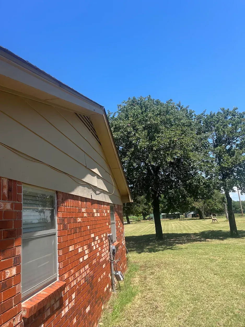 Red brick house exterior with beige siding, window, and trees under a clear blue sky.