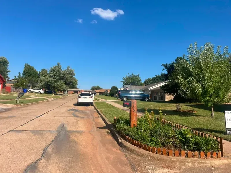 A residential street with a white car parked, green lawns, houses, and a blue sky.