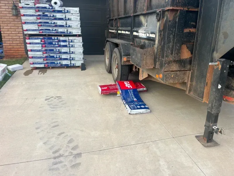 Construction materials and trailer on concrete driveway. Blue and red bags under trailer. Stack of materials to left.