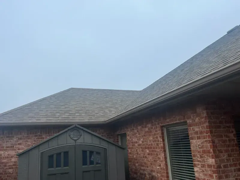 Red brick house with brown shingle roof, gray shed, and cloudy sky.