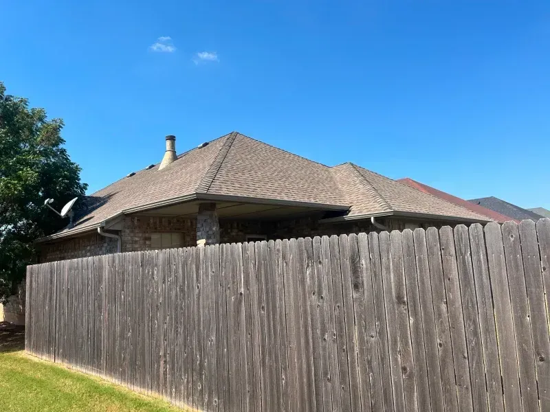 Brown shingled roof of a house with a wooden fence in front, under a clear blue sky.