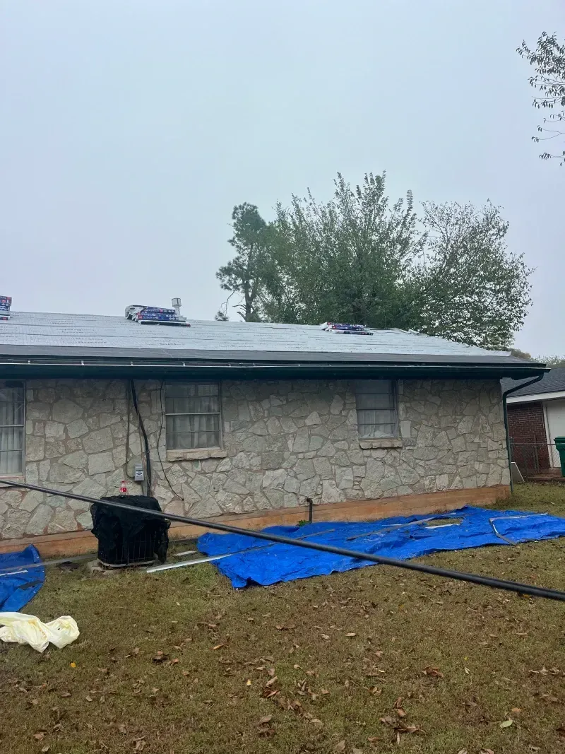 House with damaged roof covered by blue tarp, overcast day.