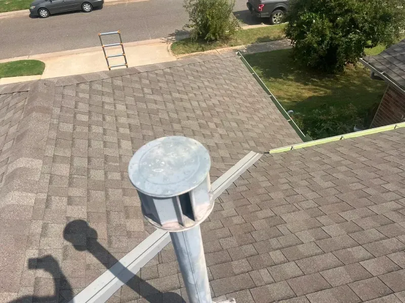 View of a roof with a silver siren. Brown shingles, gray gutters, and a street view are in the background.