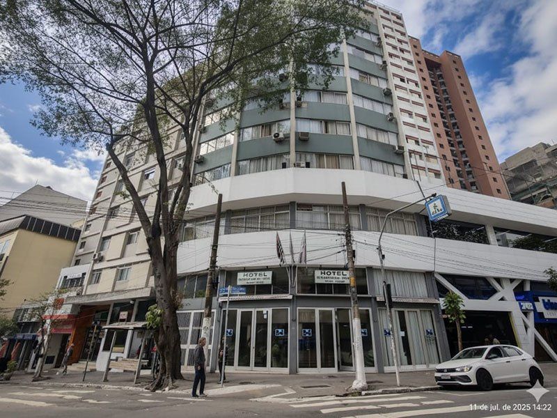 Vista de baixo para cima de um prédio de hotel de vários andares com fachada branca e janelas com tonalidade cinza em um dia ensolarado.