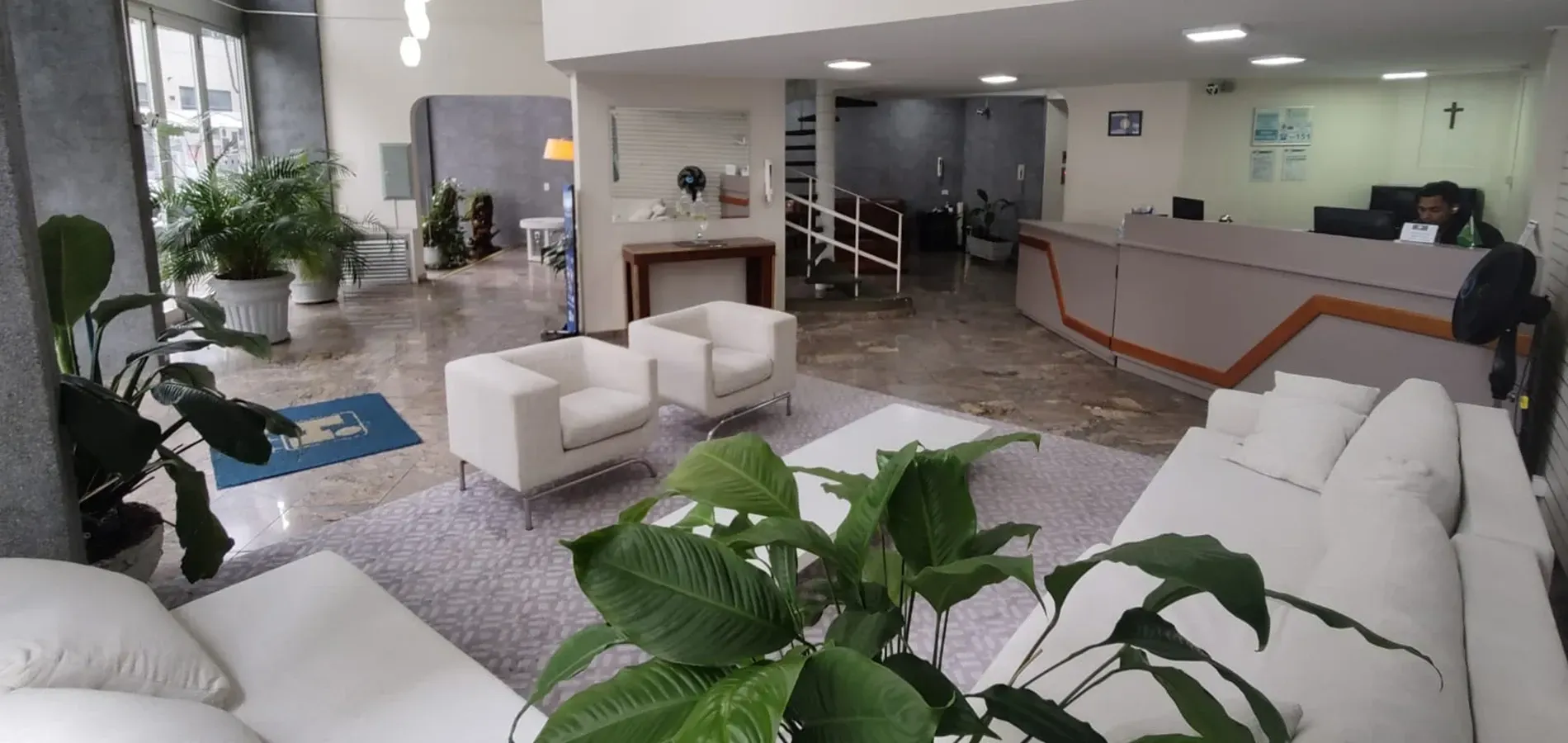 Um hall de entrada de escritório moderno e bem iluminado, com assentos brancos, balcão de recepção, piso de mármore e diversas plantas de interior.