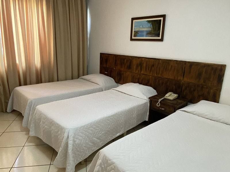 Um quarto de hotel com três camas de solteiro com roupa de cama branca, cabeceira de madeira, piso de azulejo e cortinas bege.
