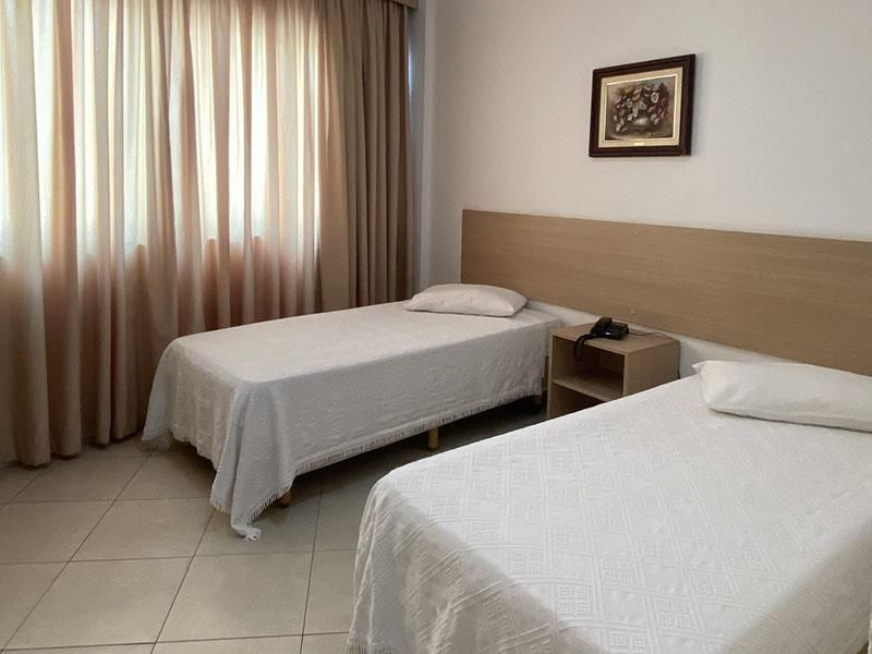 Um quarto de hotel com duas camas de solteiro brancas, uma mesa de cabeceira entre elas e cortinas bege cobrindo uma janela.
