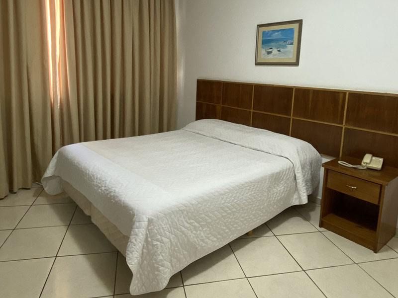 Um quarto de hotel com uma cama de casal com colchas brancas, cabeceira de madeira, uma pequena mesa de cabeceira e cortinas bege-claras.