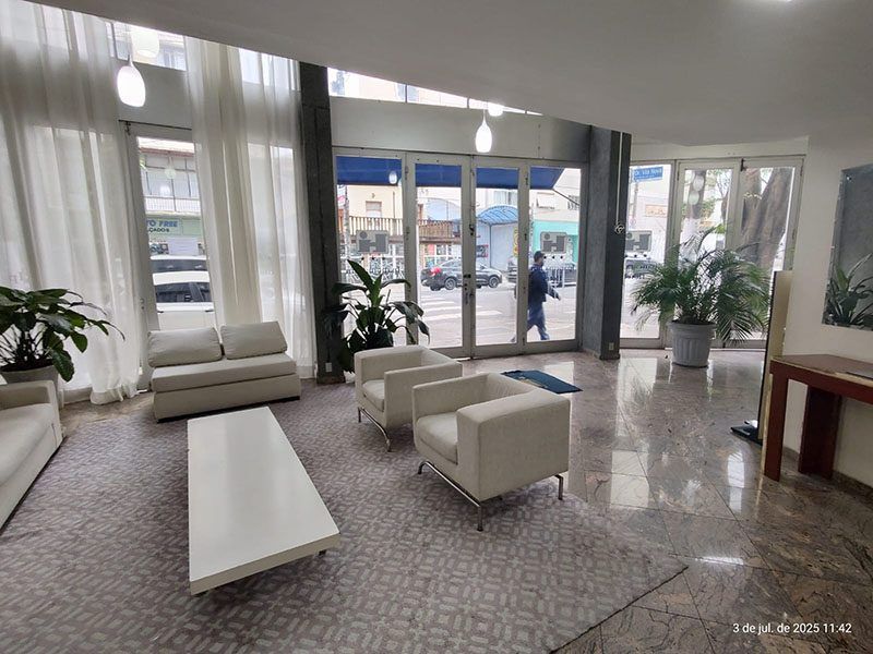 Um lobby de hotel ou escritório moderno e iluminado pelo sol, com sofás brancos, uma mesa de centro e grandes portas de entrada de vidro.