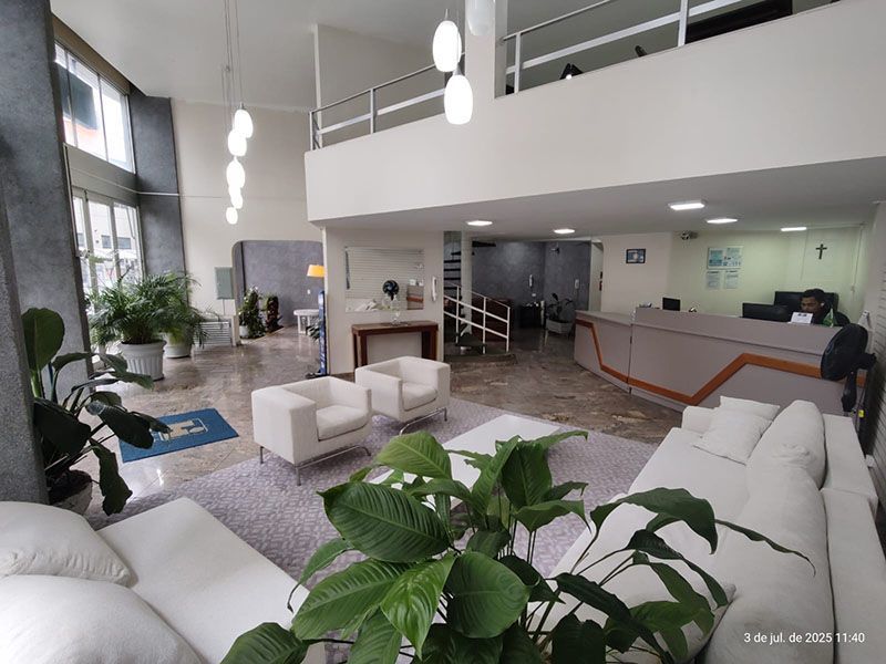 Um saguão de hotel luminoso com poltronas e sofás brancos, plantas de interior e um balcão de recepção ao fundo.
