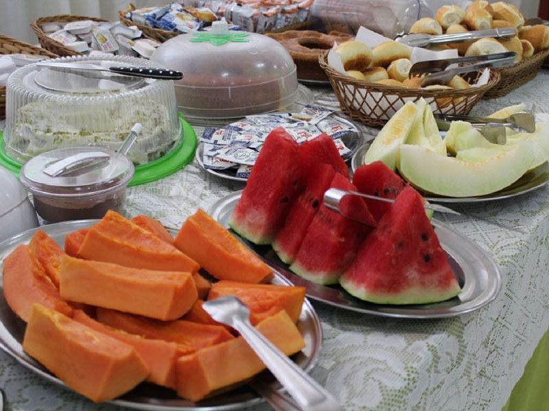 Uma mesa de bufê com bandejas de mamão papaia, melancia e melão fatiados, ao lado de cestas de pão e pequenos recipientes com alimentos.