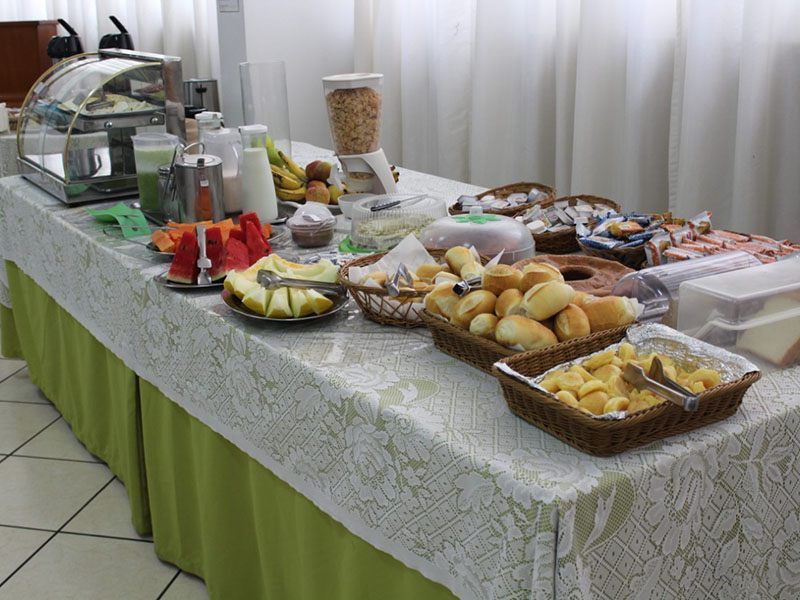 Uma mesa de buffet de café da manhã com diversos tipos de pães, frutas fatiadas, suco e cereais sobre uma toalha de mesa branca com saia verde.