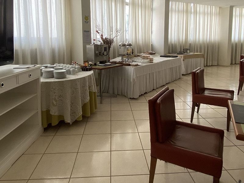 Uma sala de jantar com mesas redondas e compridas cobertas com toalhas brancas, preparada para uma refeição perto de grandes janelas com cortinas transparentes.