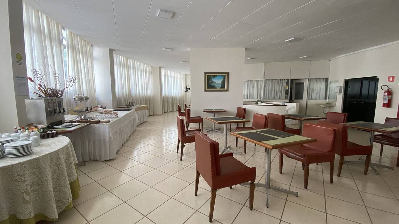 Sala de café da manhã de hotel com mesa de bufê, mesas de jantar com cadeiras vermelhas, piso de azulejo e cortinas brancas.