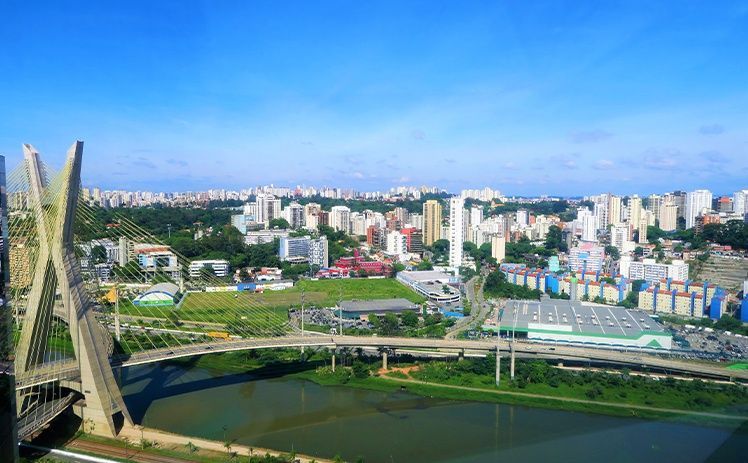 Uma vista ensolarada e de cima da ponte estaiada Octávio Frias de Oliveira sobre um rio, com o horizonte da cidade ao fundo.