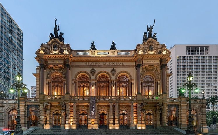 O Teatro Municipal de São Paulo é um edifício bege, ornamentado em estilo Beaux-Arts, que ao entardecer é iluminado por altas estátuas.