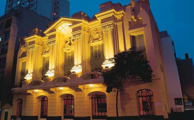 Um edifício neoclássico histórico e iluminado ao entardecer, com colunas ornamentadas e grandes janelas em arco.