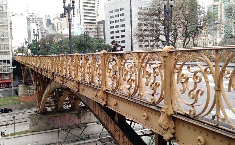 Uma ponte ornamentada de metal amarelo com grades circulares em forma de arabescos, em um ambiente urbano, vista de um ângulo ligeiramente elevado.
