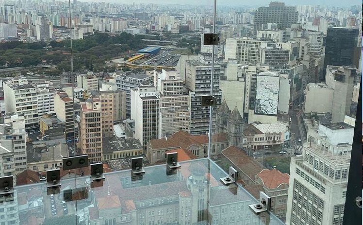 Vista de cima, através de um piso de vidro, de uma paisagem urbana densa e cinzenta, com prédios altos e ruas movimentadas.
