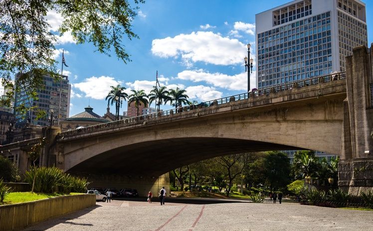 Uma ponte arqueada de pedra atravessa um parque com palmeiras sob um céu azul brilhante, com edifícios altos ao fundo.