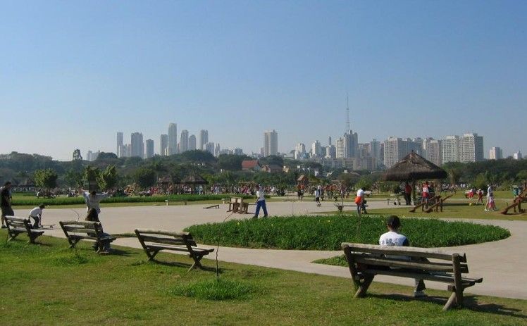 Pessoas relaxam e brincam em um parque com bancos e um campo gramado, tendo como pano de fundo uma paisagem urbana distante sob um céu azul.