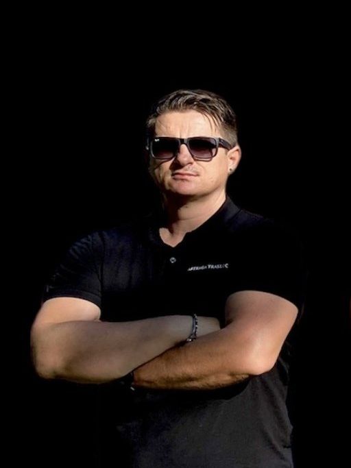 Un uomo che indossa occhiali da sole e una camicia nera è in piedi con le braccia incrociate. Sem del team Arteaga