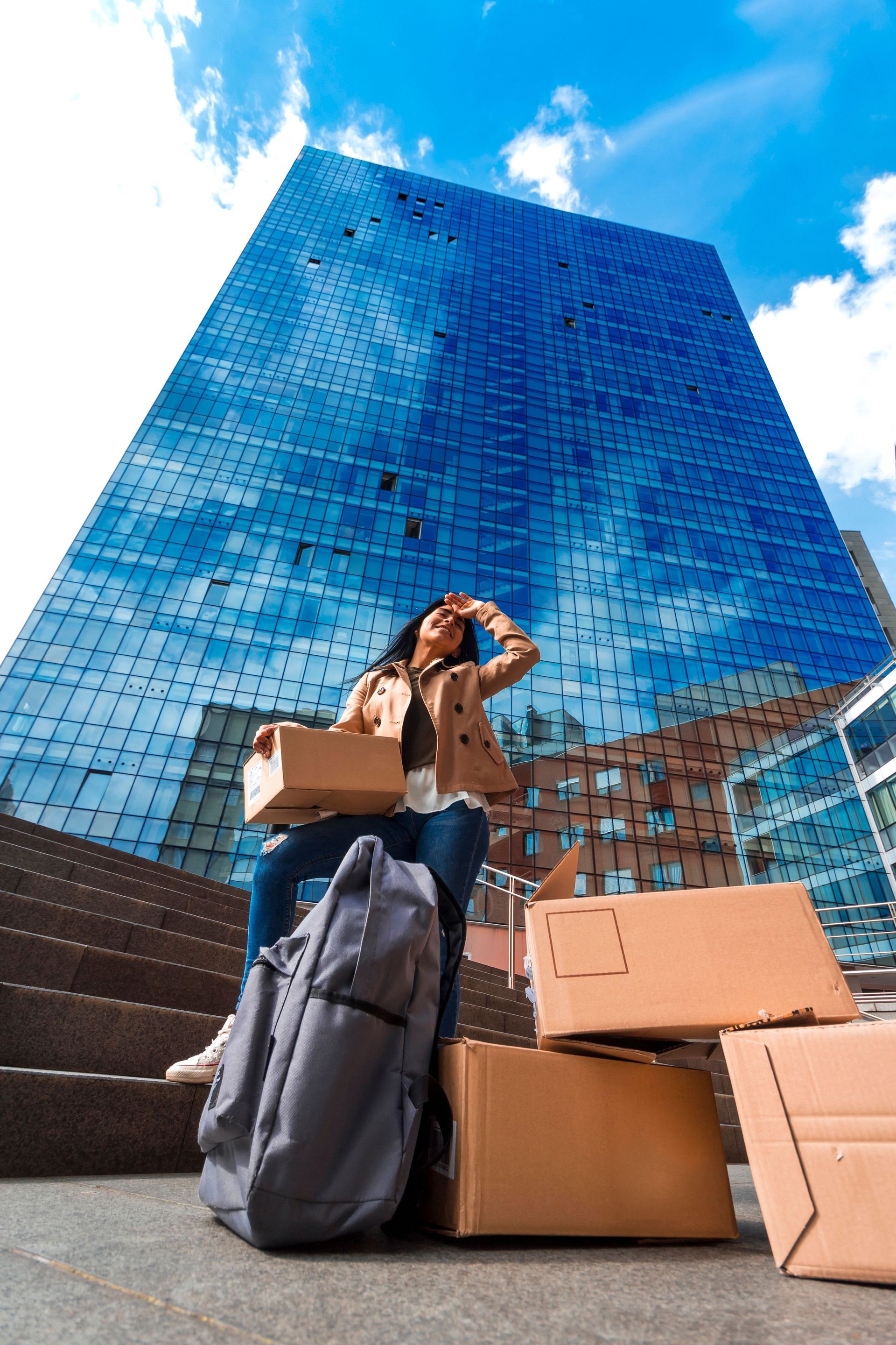Person with boxes and backpack standing before a tall blue glass building under a bright sky