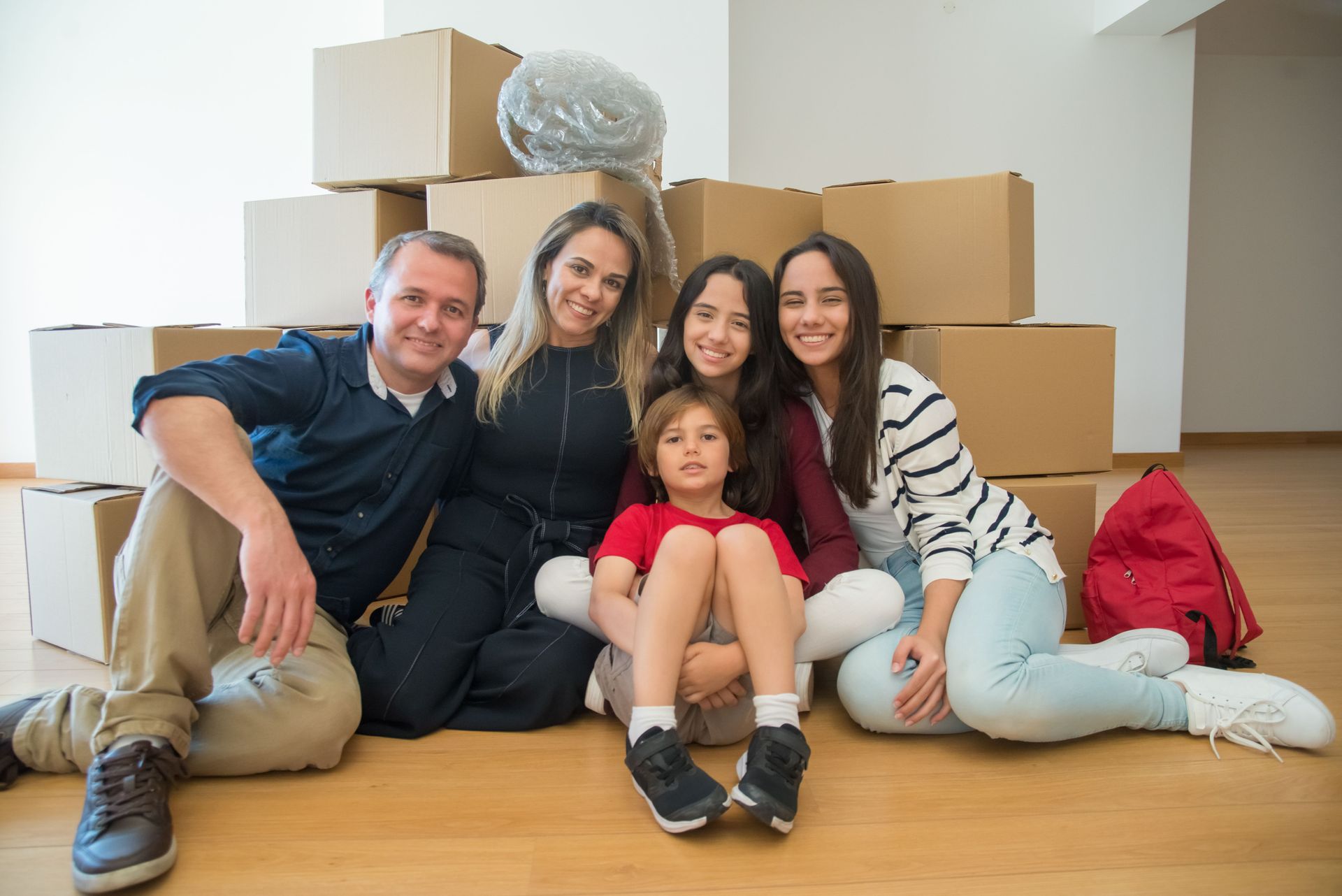 Family sitting on the floor in a new home, surrounded by stacked moving boxes, smiling at the camera.