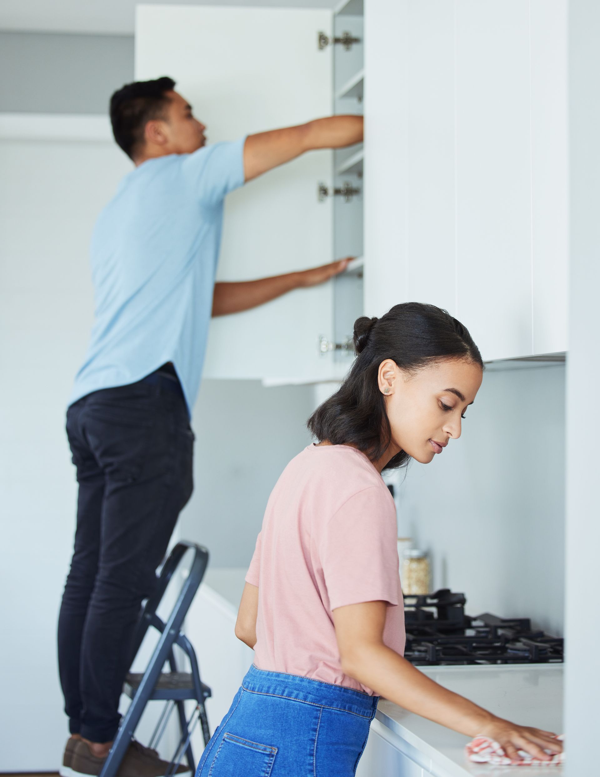 A man is standing on a ladder while a woman is cleaning a kitchen counter.