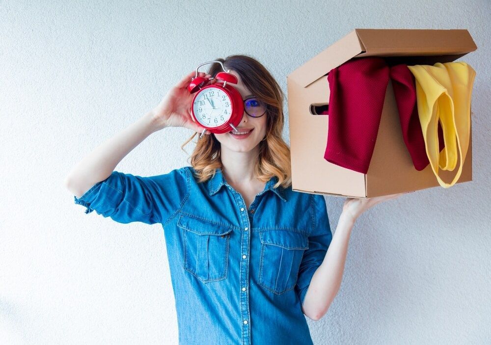 A woman is holding a box of clothes and an alarm clock in front of her face.