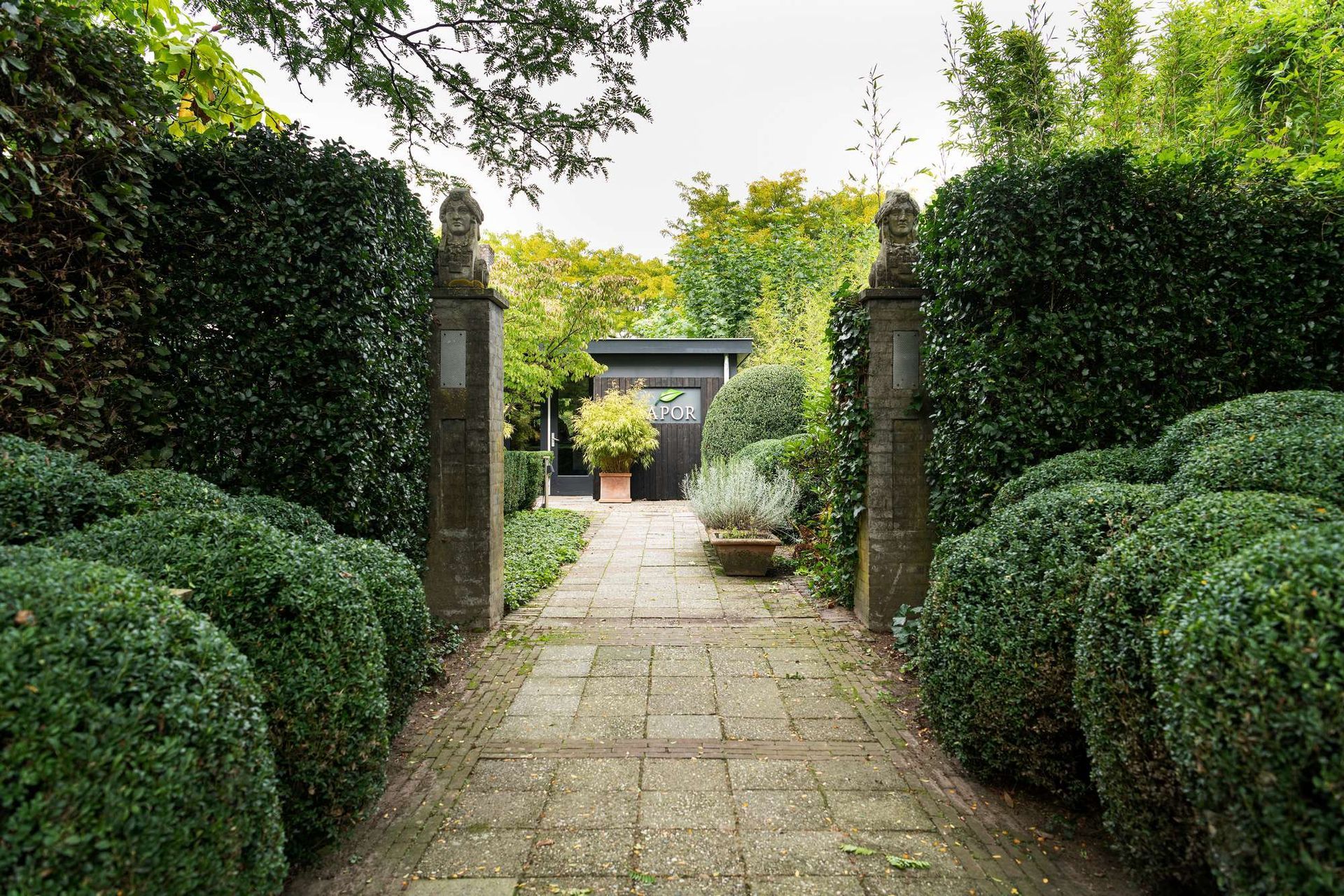 Een stenen pad door een formele tuin met strak gesnoeide groene hagen leidt naar een donker gebouw.
