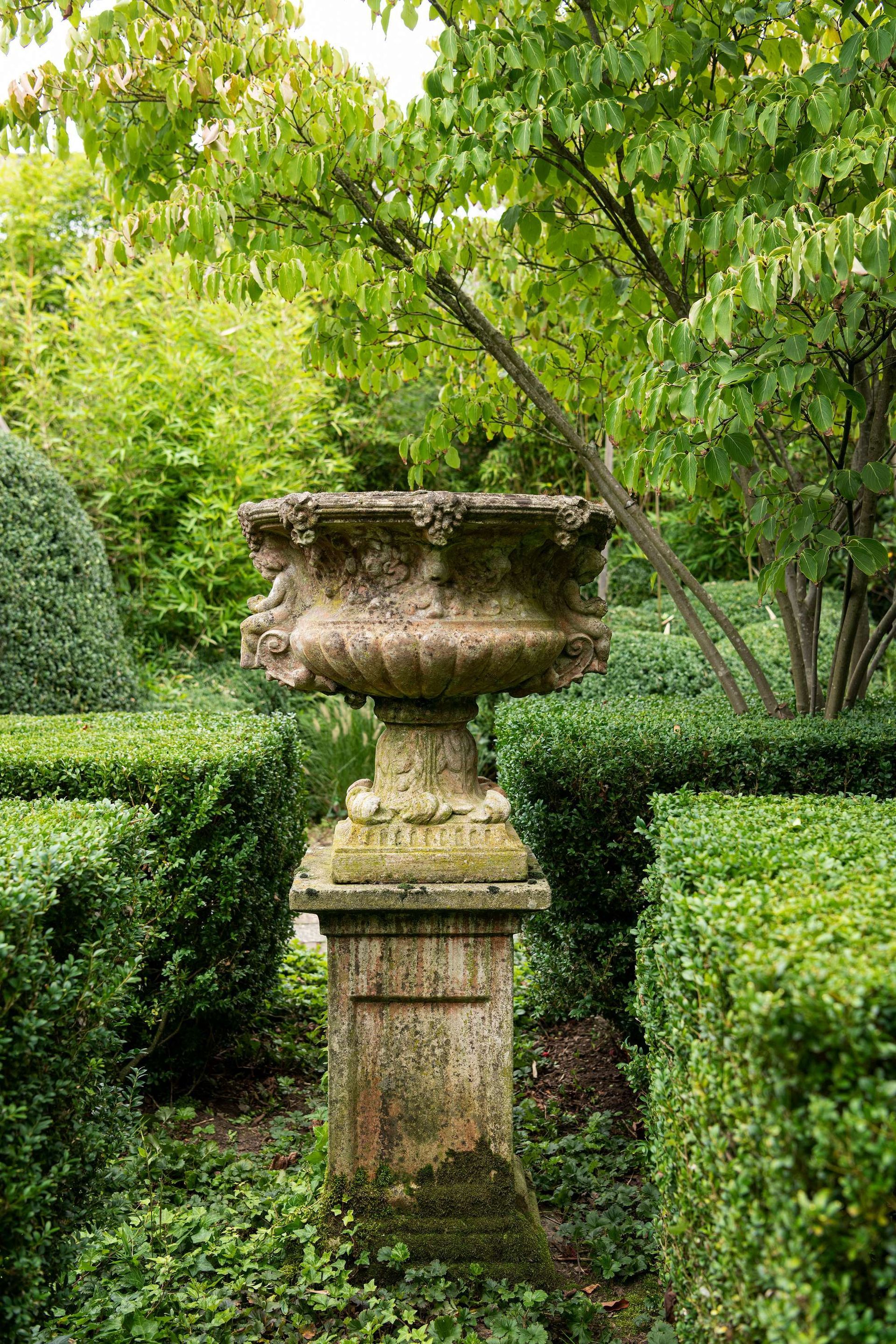 Een verweerde stenen plantenbak op een sokkel in een formele tuin, omgeven door keurig gesnoeide hagen.
